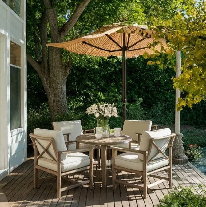 A outdoor patio with a round wooden table, four chairs with white cushions, and a beige umbrella for shade.